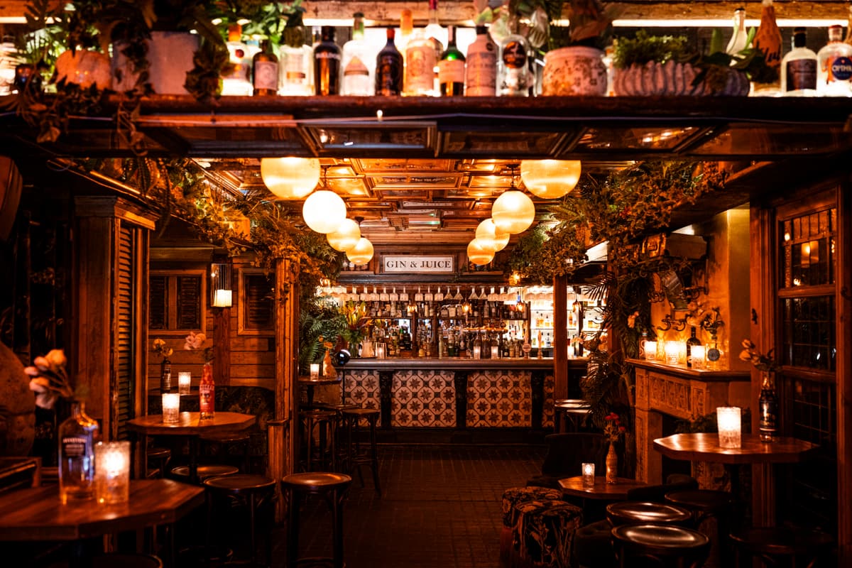 Dimly lit bar with globe pendant lights, shelves of bottles overhead, patterned tiles on the bar front and candlelit wooden tables.
